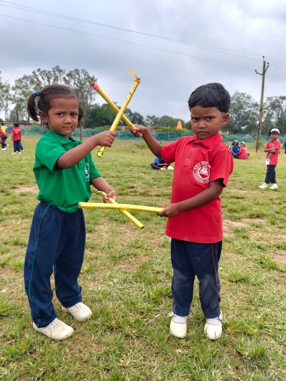 Two young pupils in green and red house shirts playing with yellow sticks on the ground