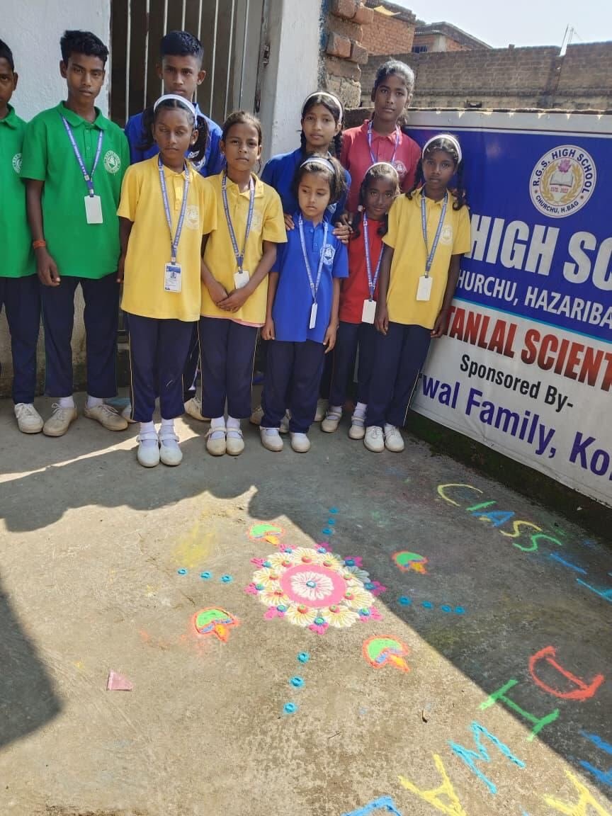 Students in house shirts standing beside a rangoli and the RGS school banner